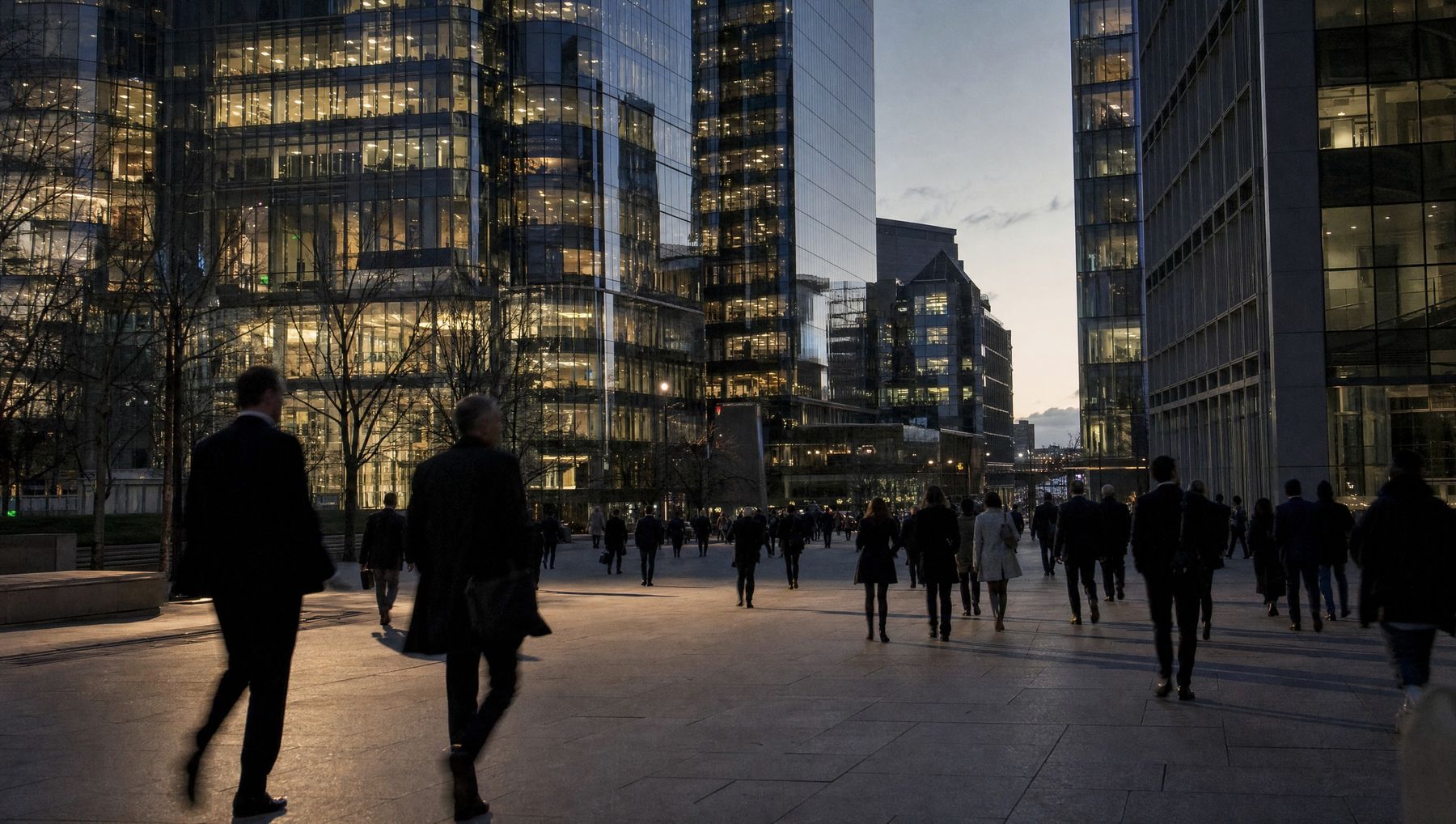 Office workers in coats crossing a plaza between lit glass towers at dusk.